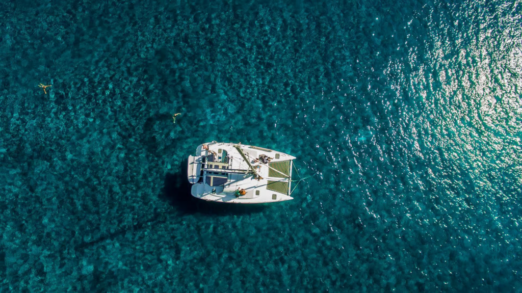 private boat tour Santorini aerial view of luxury catamaran floating above crystal-clear turquoise water, highlighting an exclusive and scenic island sailing experience