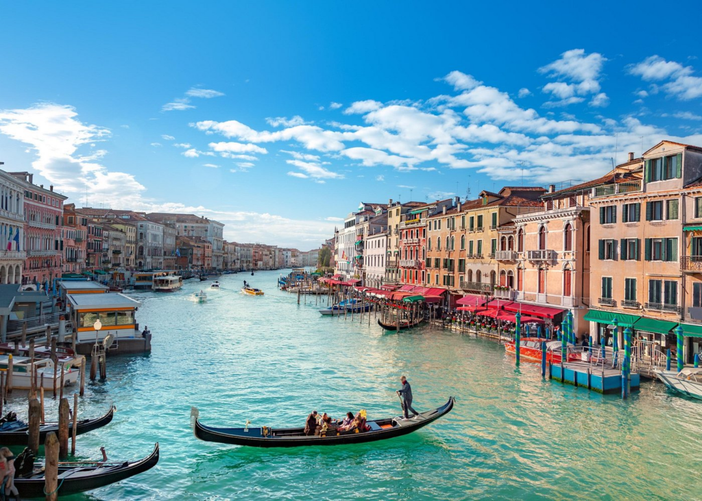 Scenic view of a gondola traveling along Venice’s Grand Canal with historic waterfront buildings and lively streets under a bright blue sky, capturing the iconic setting that complements experiences on Venice Food And Wine Tours.