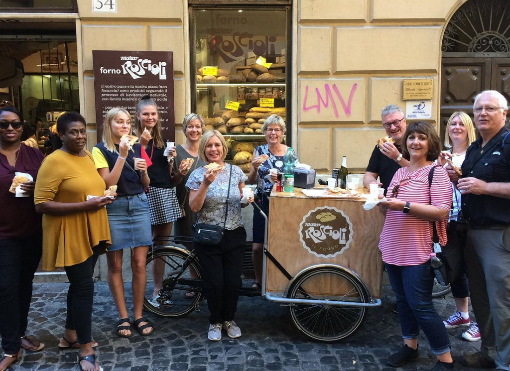 Tour group tasting fresh pizza and bread during rome market tours outside a local bakery in Rome