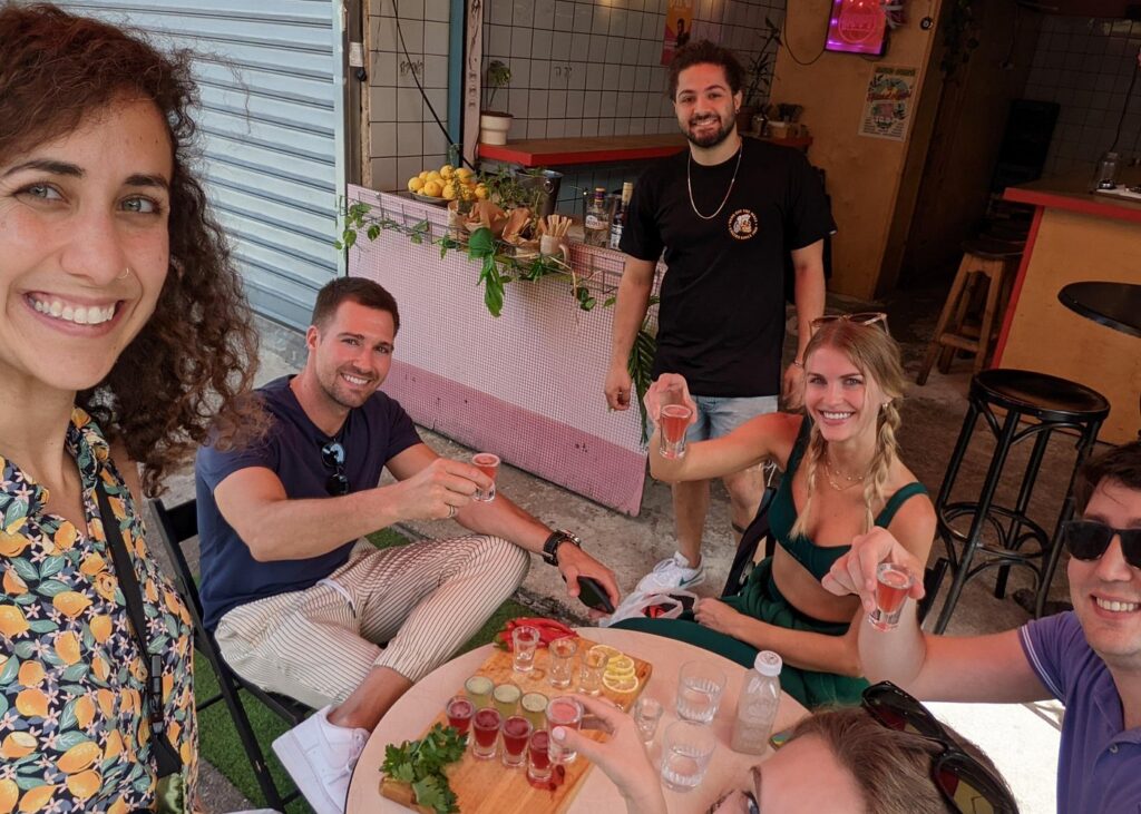 Carmel Market Tel Aviv food tour with travelers enjoying fresh fruit shots and local tastings at a vibrant market cafΓ© surrounded by vendors and neighborhood eateries.