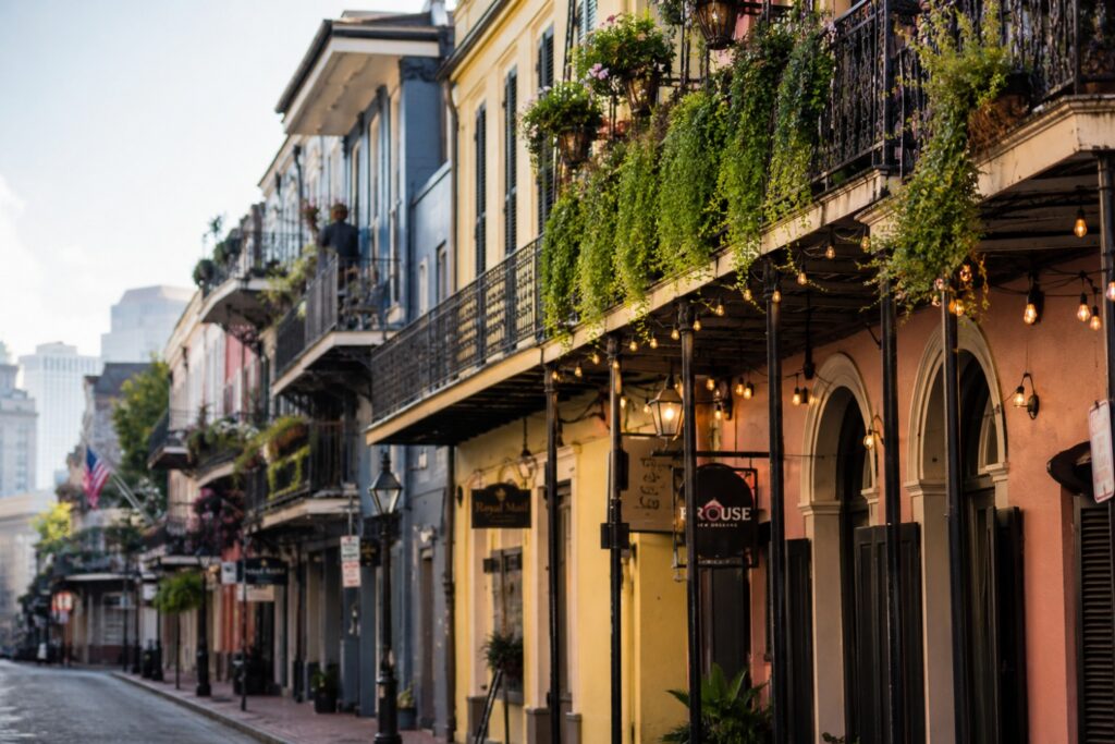 A wide daytime photograph of a French Quarter street in New Orleans, wrought iron balconies dripping with ferns and string lights, pastel colored Creole townhouses in pink, yellow and blue, cobblestone street, soft afternoon light, warm inviting travel photography style, no visible people's faces, shallow depth of field on the balconies in the foreground.