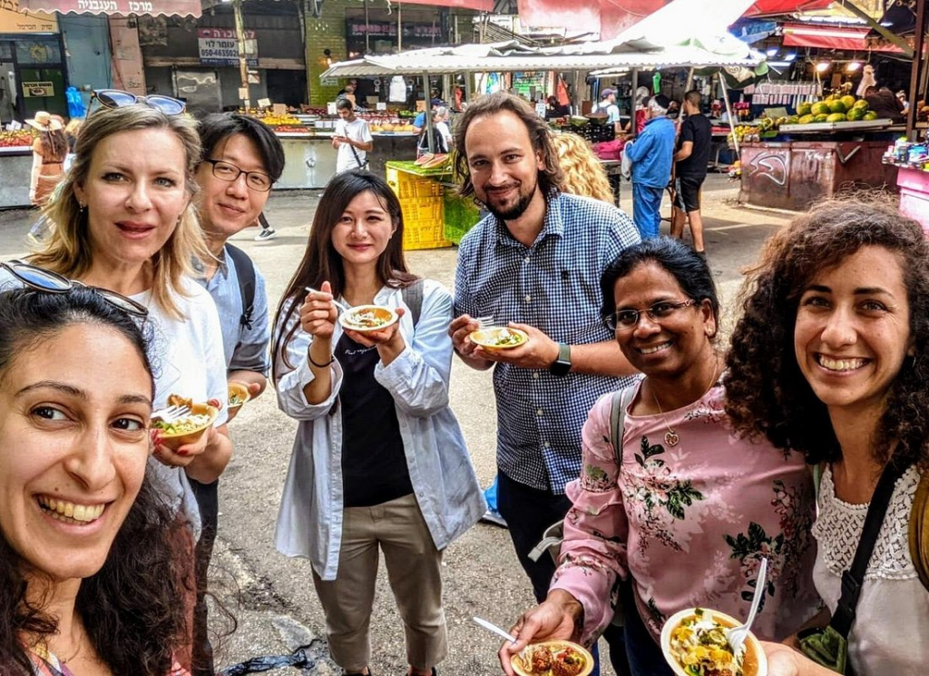 Carmel Market Tel Aviv food tour with a diverse group of travelers sampling authentic Israeli street food while exploring the bustling open-air market with a local guide.