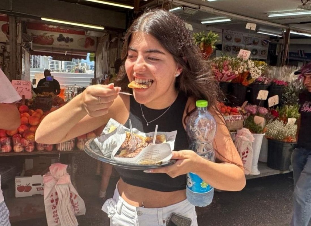Carmel Market Tel Aviv featuring a traveler enjoying freshly prepared Israeli street food while exploring the vibrant market filled with fresh produce, flowers, and local vendors.