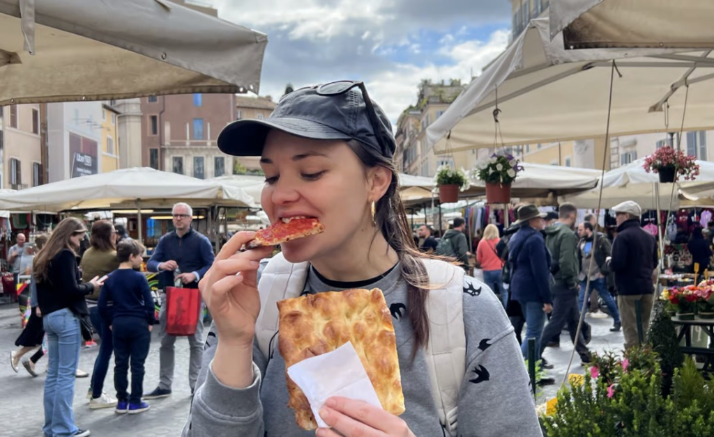 Rome food and wine tours featuring a traveler enjoying authentic Roman pizza by the slice while exploring a bustling outdoor market in the heart of Rome.
