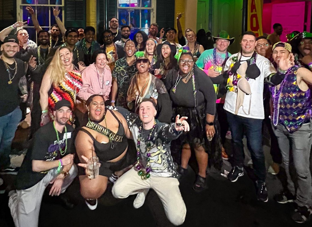 Large group of friends wearing Mardi Gras beads and costumes during a new orleans pub crawl, posing outside lively bars in the French Quarter at night.