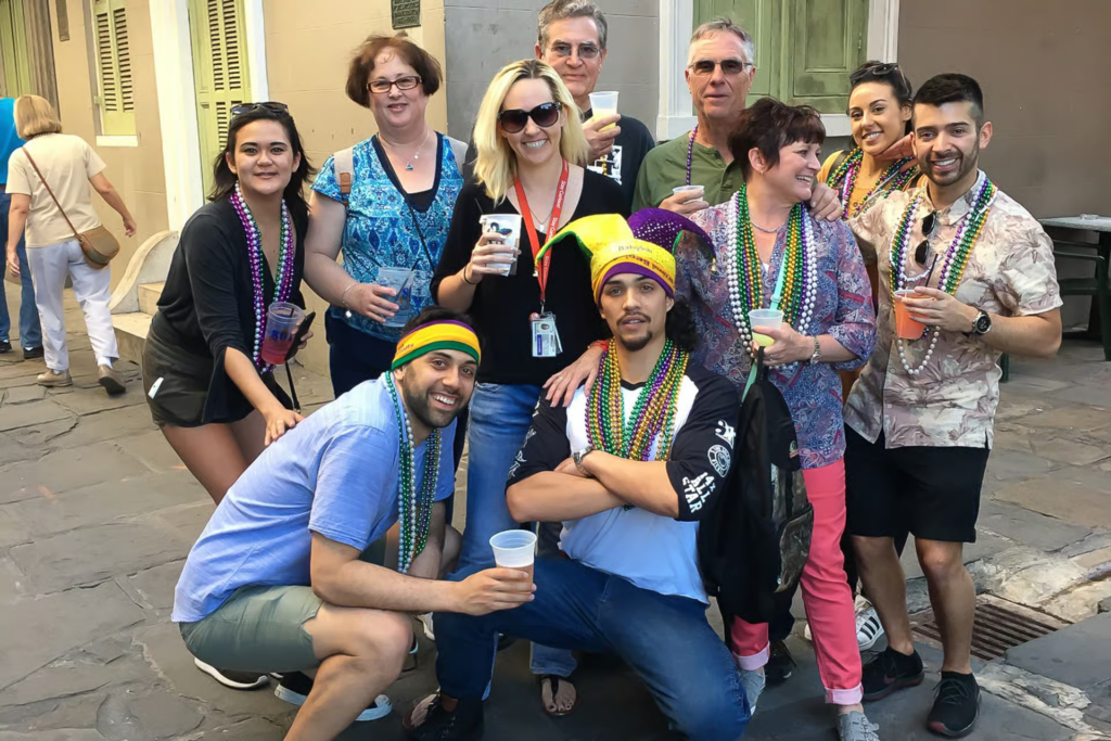 Group of adults wearing Mardi Gras beads and holding drinks during a new orleans pub crawl, gathered together on a French Quarter street.
