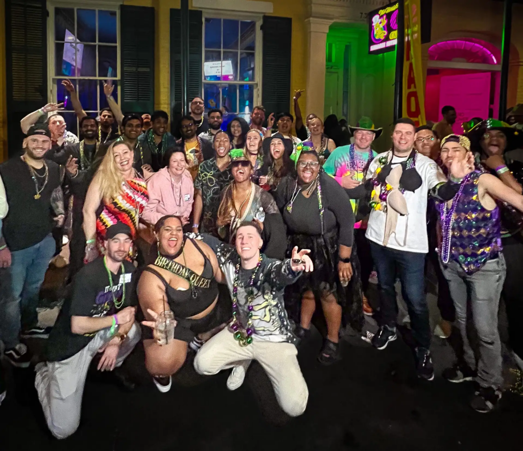 Large group celebrating outside French Quarter bars at night during a new orleans pub crawl, wearing Mardi Gras beads and festive costumes.