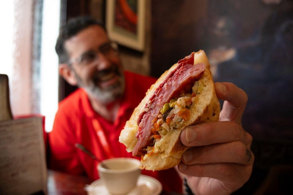 A guest holds up a classic New Orleans muffuletta sandwich filled with layers of Italian meats, cheese, and olive salad during a French Quarter food tour.