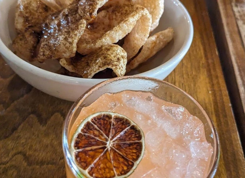 A handcrafted cocktail garnished with dried citrus is served alongside crispy pork cracklings during a French Quarter food tour, highlighting classic New Orleans flavors and local bar bites.